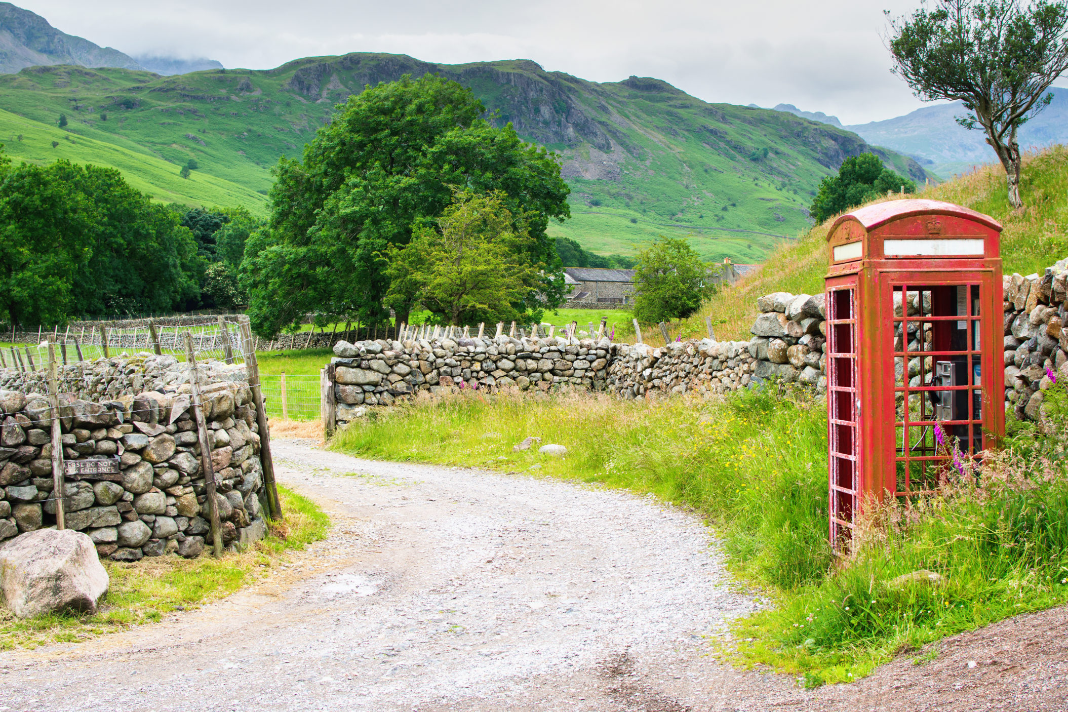 A narrow, gravel road winds between stone walls. On the side of the road is a red phone booth. Trees, green fields, and mountains are all around. [Shutterstock]