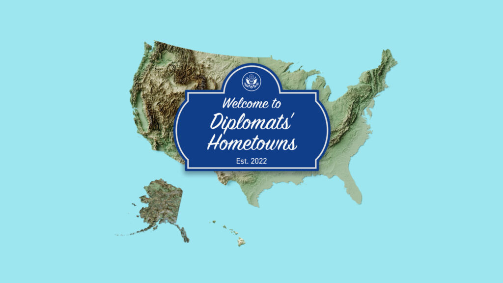 topographic map of United States over a light blue background, in foreground a dark blue sign with white text reads "Welcome to Diplomats' Hometowns Est. 2022"