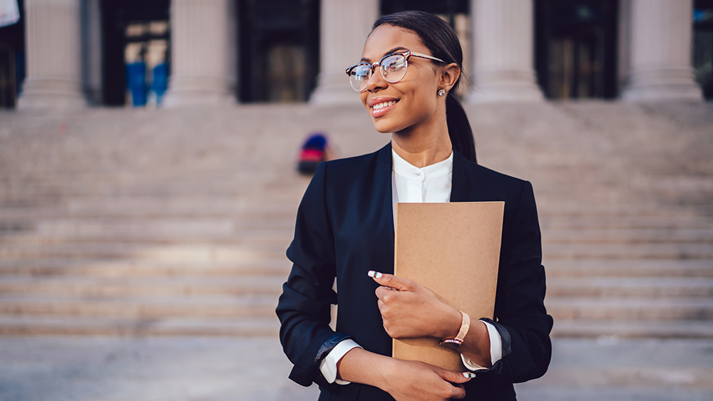 Successful African American graduate juridical specialist standing outdoors against courthouse building with folder in hands.