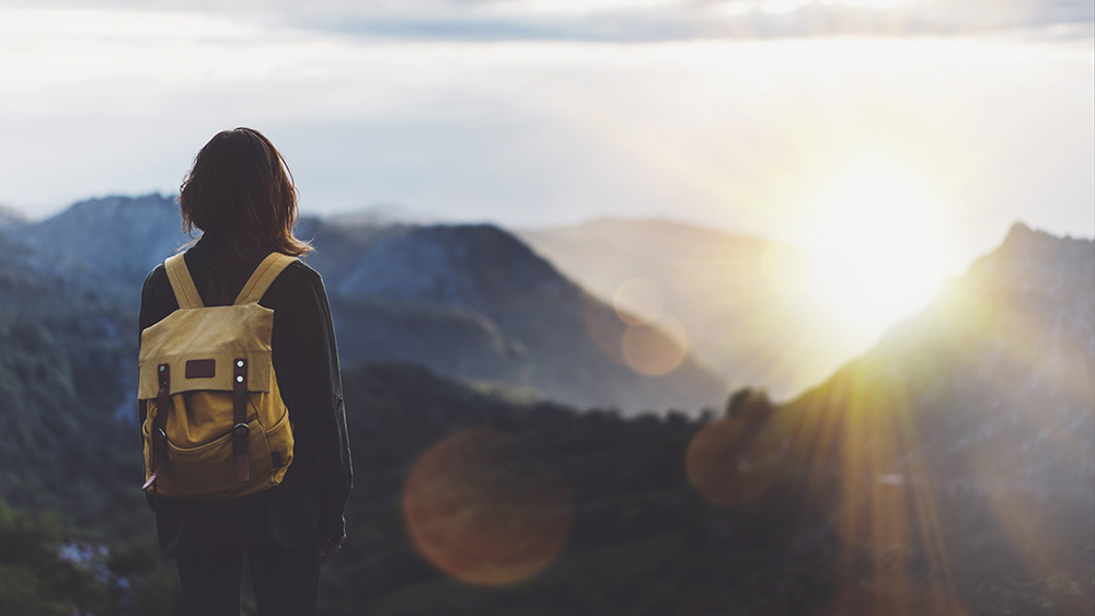 Hipster young girl with backpack enjoying sunset on peak of foggy mountain.