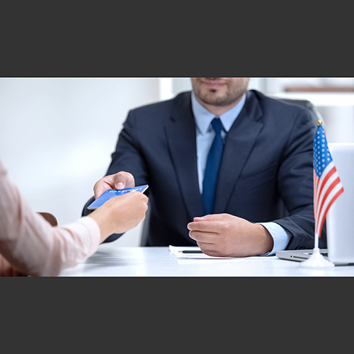 A man in a suit hands a passport to a woman. A small American flag is off to the side.